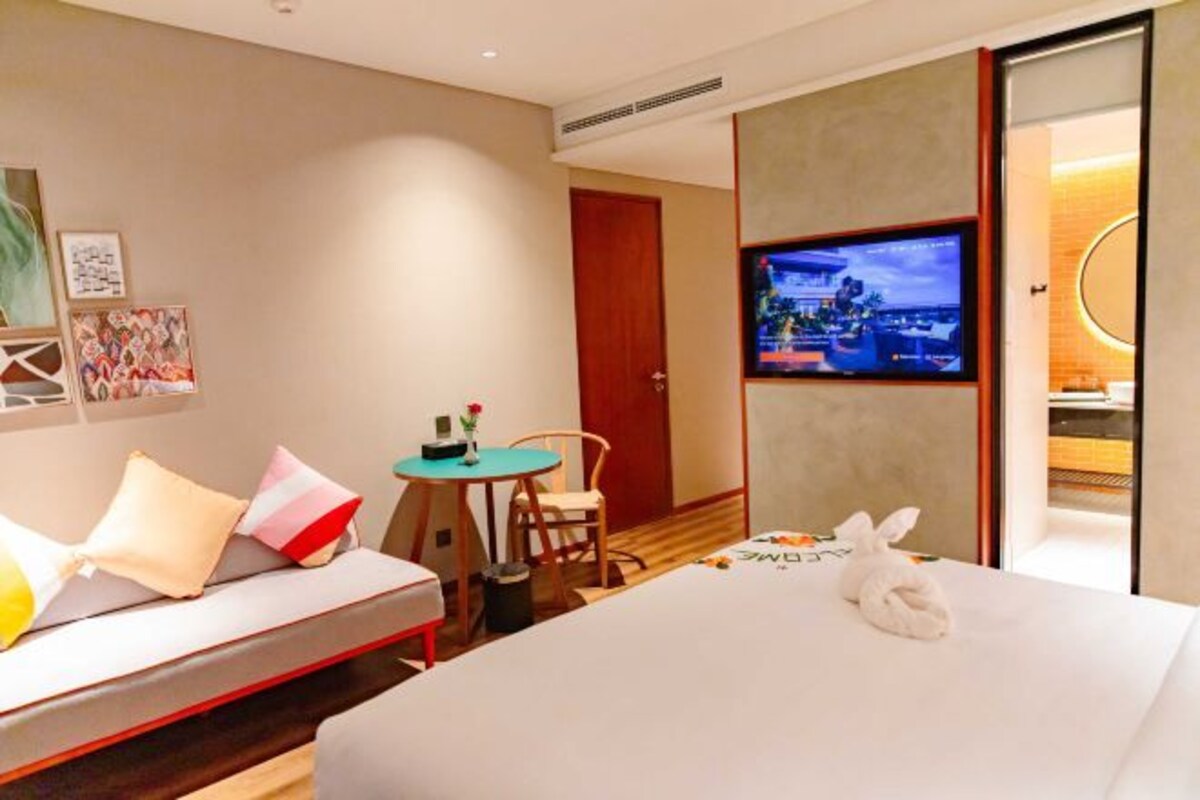 A hotel room with a bed, a small table with a chair, a TV on the wall, and artwork on the wall.