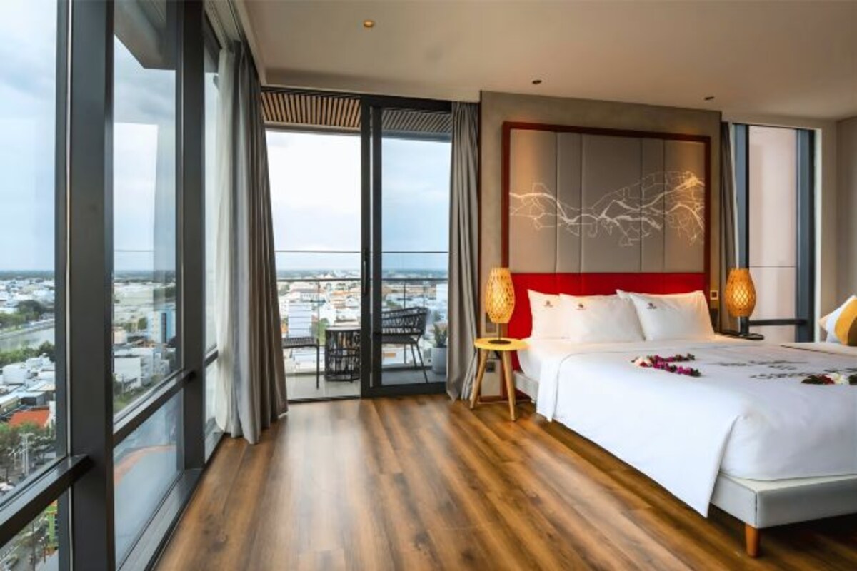 A modern hotel room with a large bed, a view of the city through floor-to-ceiling windows, and a balcony with outdoor furniture.