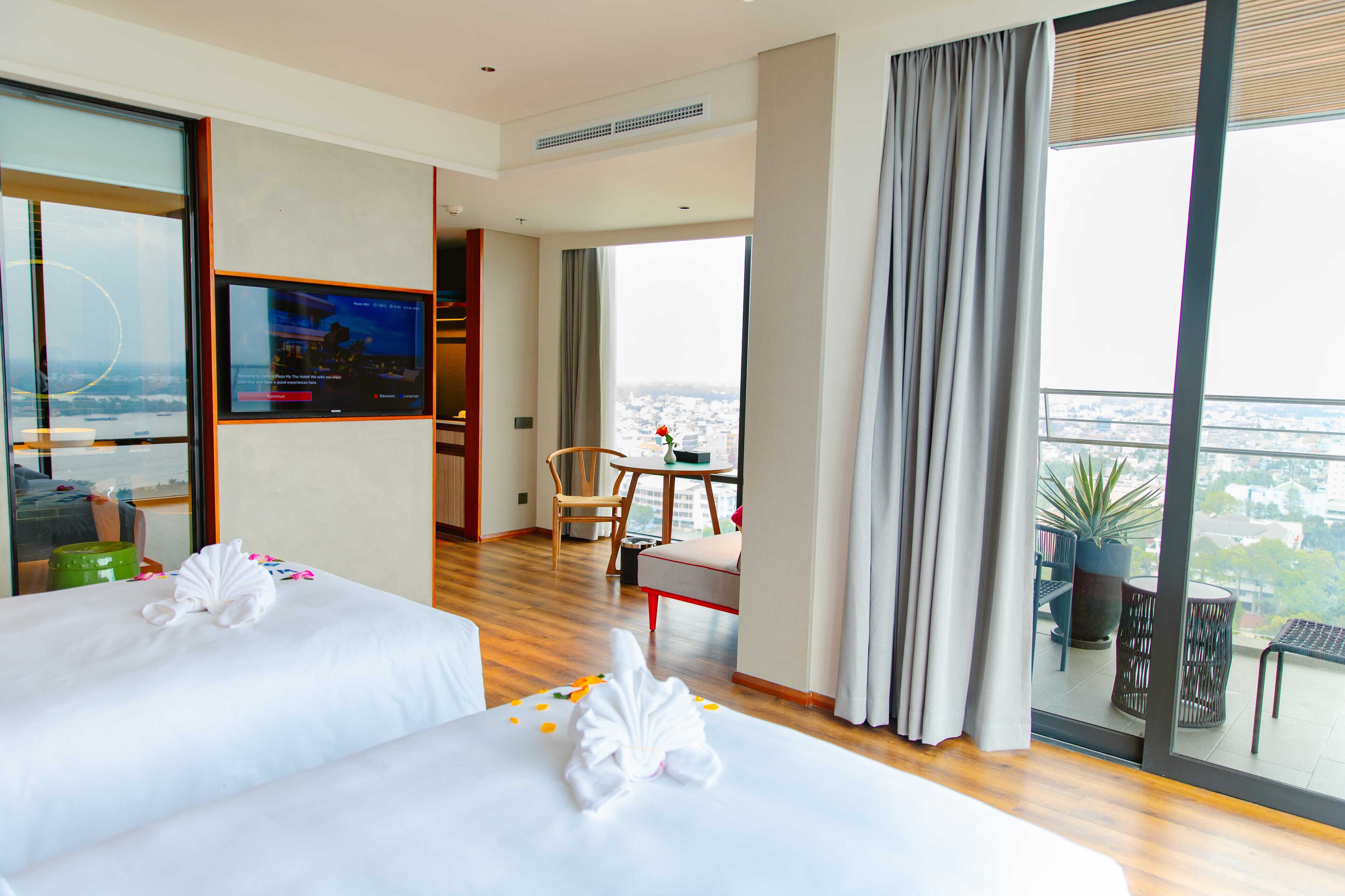 A hotel room with two beds, a large window with a city view, a balcony with plants, and a dining area with a table and chairs.
