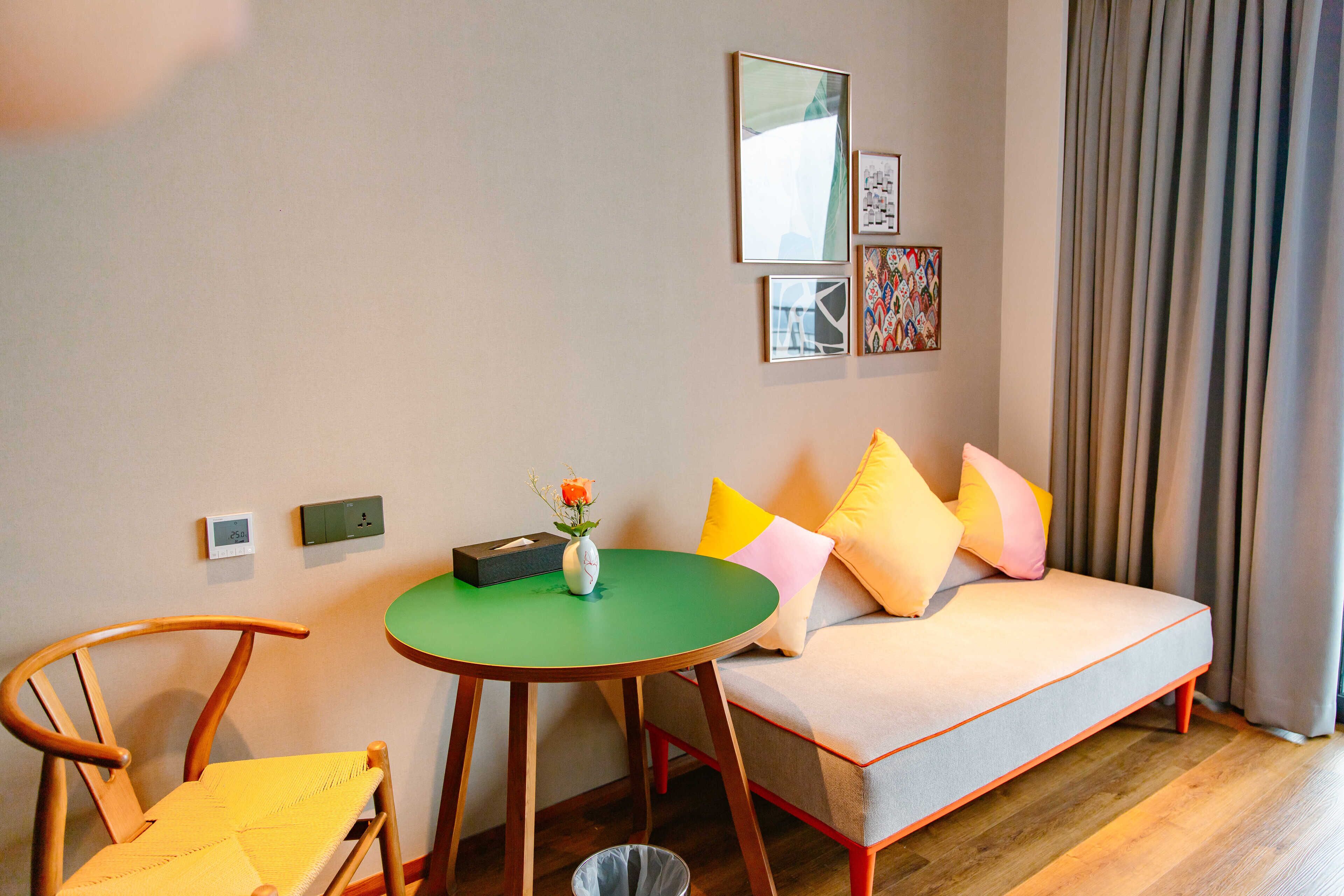 A cozy room with a green round table, a wooden chair, a sofa with colorful cushions, and a small vase with flowers.