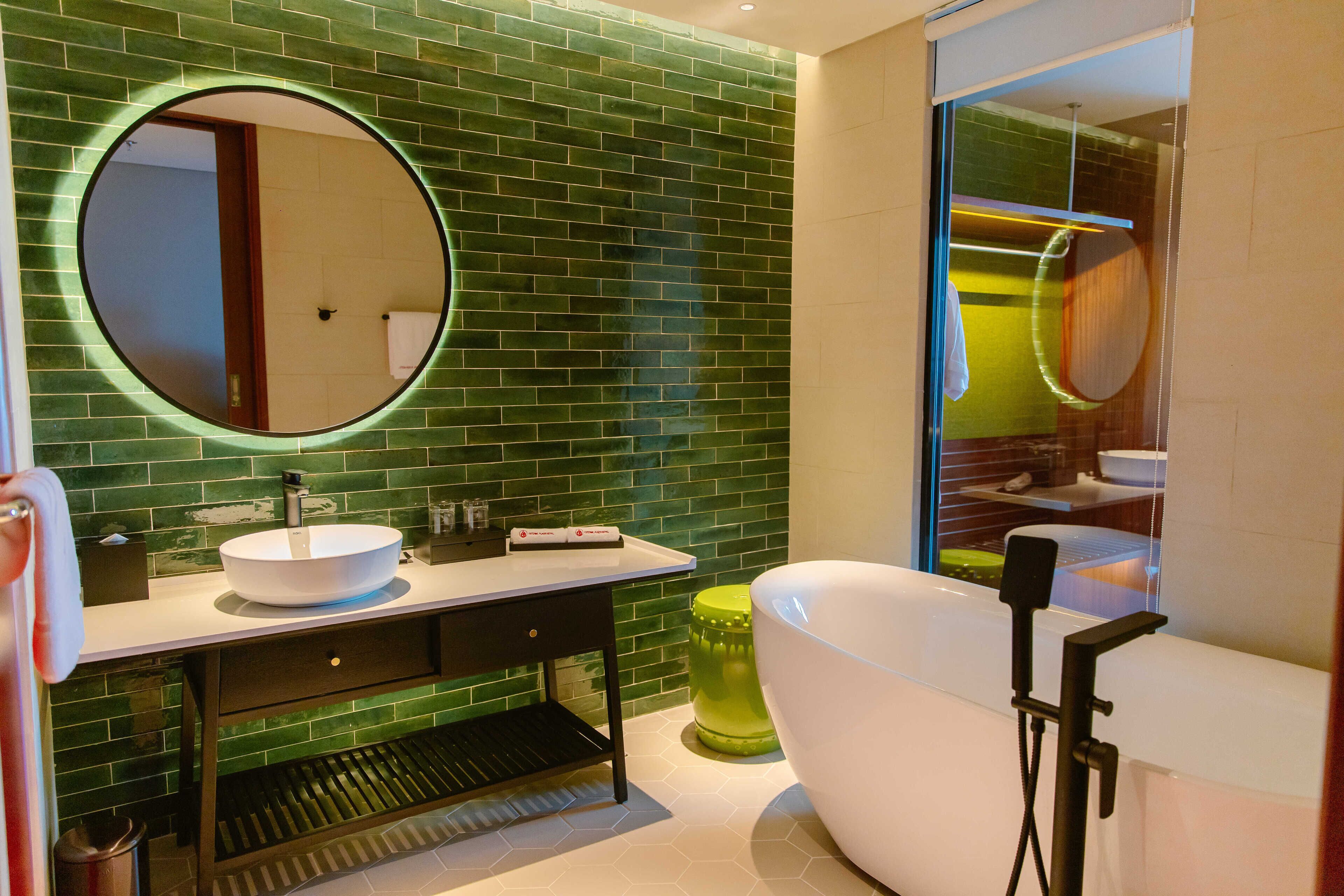 A modern bathroom with a green-tiled wall, a round mirror, a white sink, a black vanity, a green trash bin, and a white freestanding bathtub.