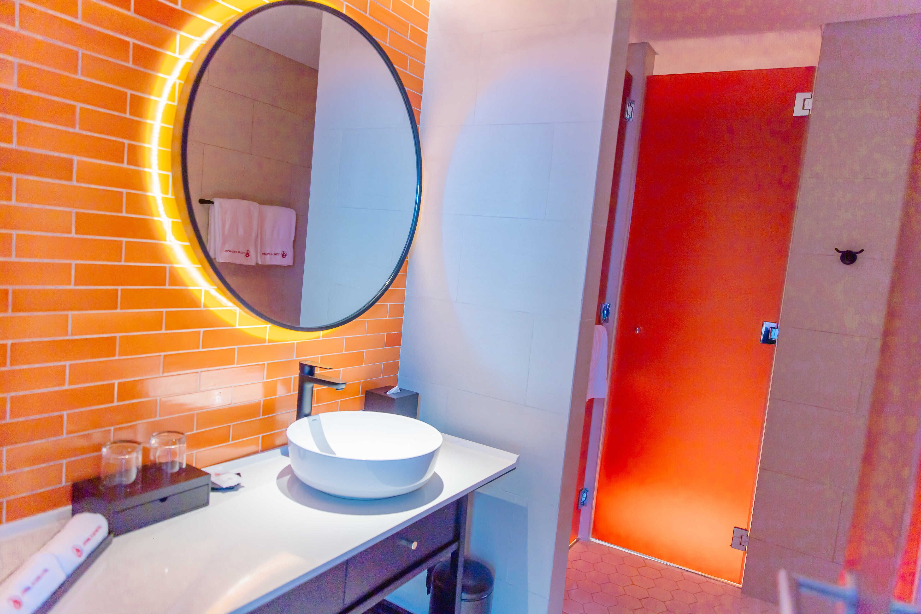 Ein Badezimmer mit rundem Spiegel, weißem Waschbecken und einer orangefarben gefliesten Wand.