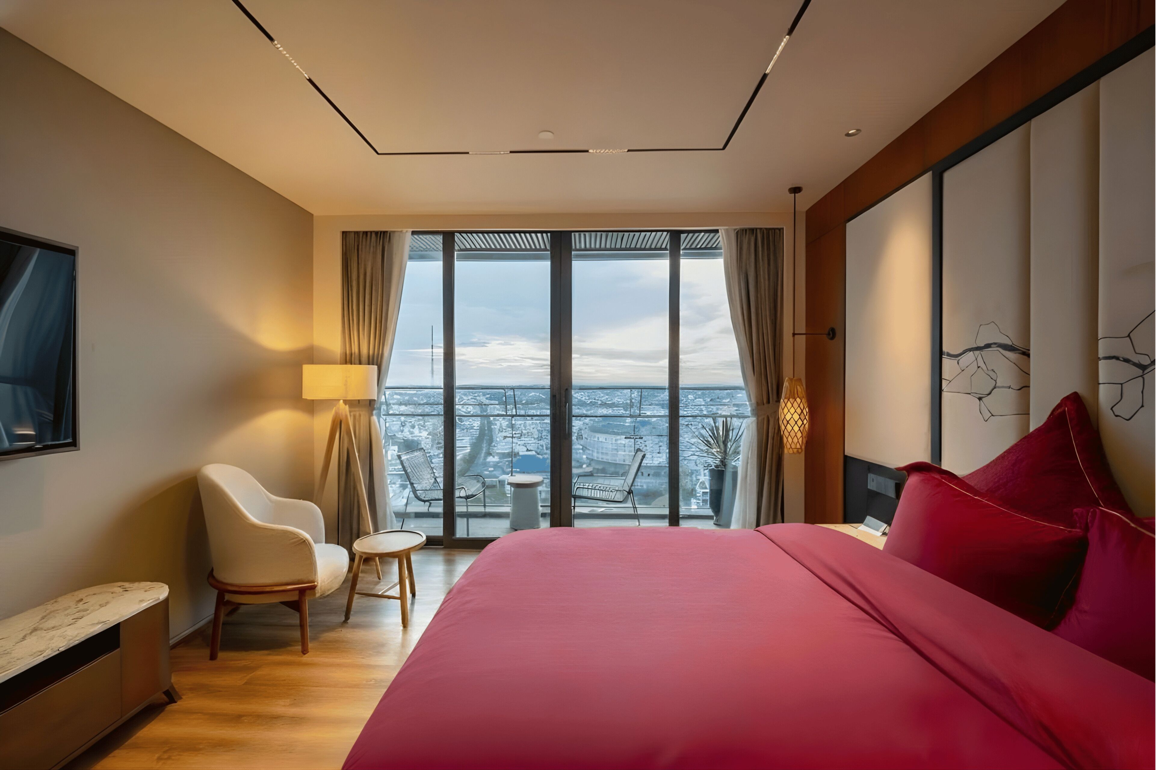 Una habitación de hotel moderna con una cama grande, un área de descanso con sillas, una mesita pequeña y un ventanal con vistas a la ciudad.