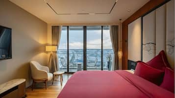 Una habitación de hotel moderna con una cama grande, un área de descanso con sillas, una mesita pequeña y un ventanal con vistas a la ciudad.