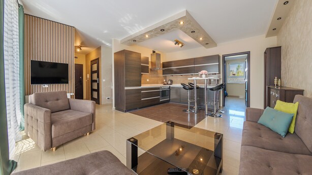 A modern living room with a sofa, a coffee table, and a TV. There is a kitchen area with a dining table and bar stools. - Apartamenty Sun & Snow Villa Nord (Wladyslawowo)