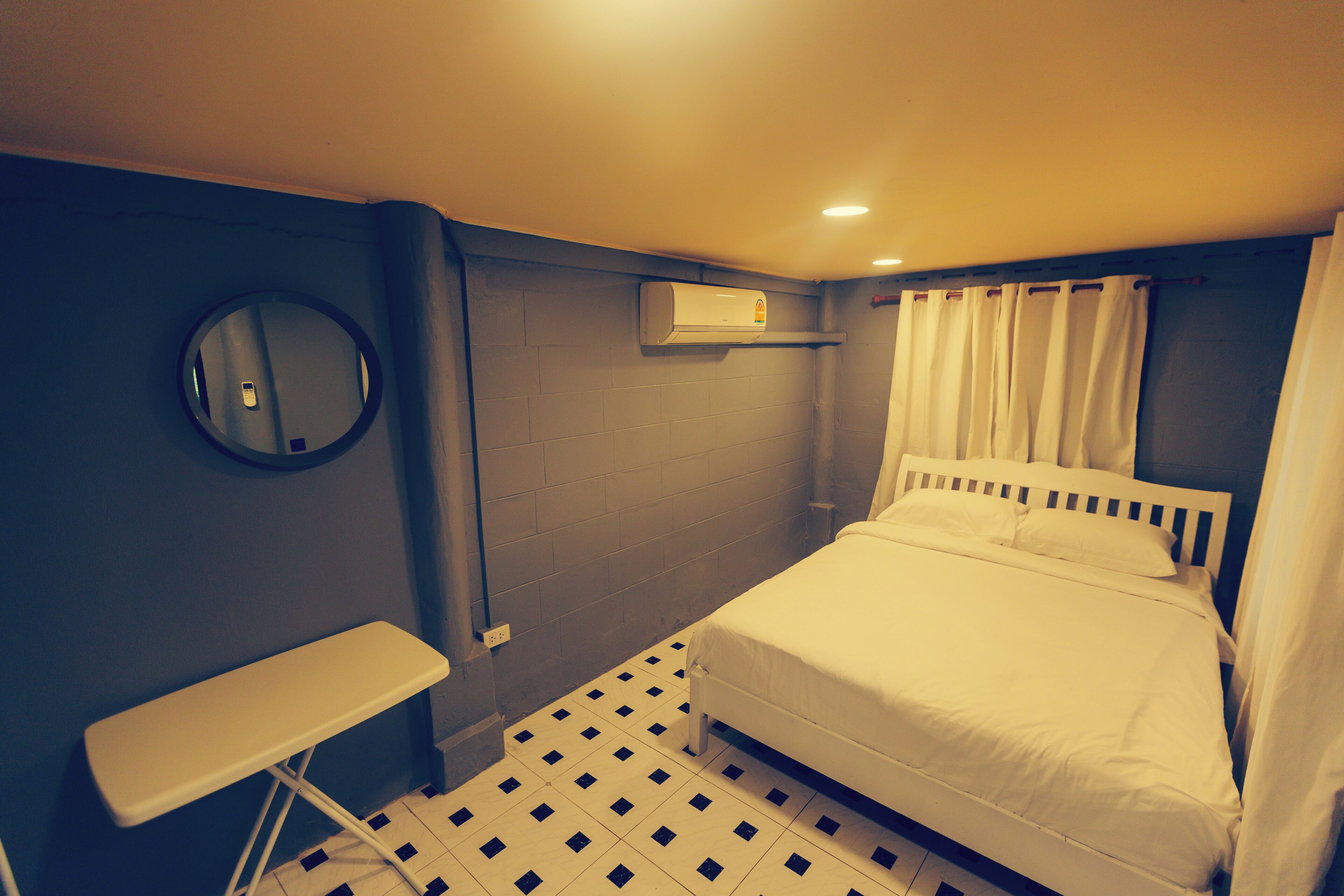 A small, compact hotel room with a single bed, a small table, a mirror, and a wall-mounted air conditioner.