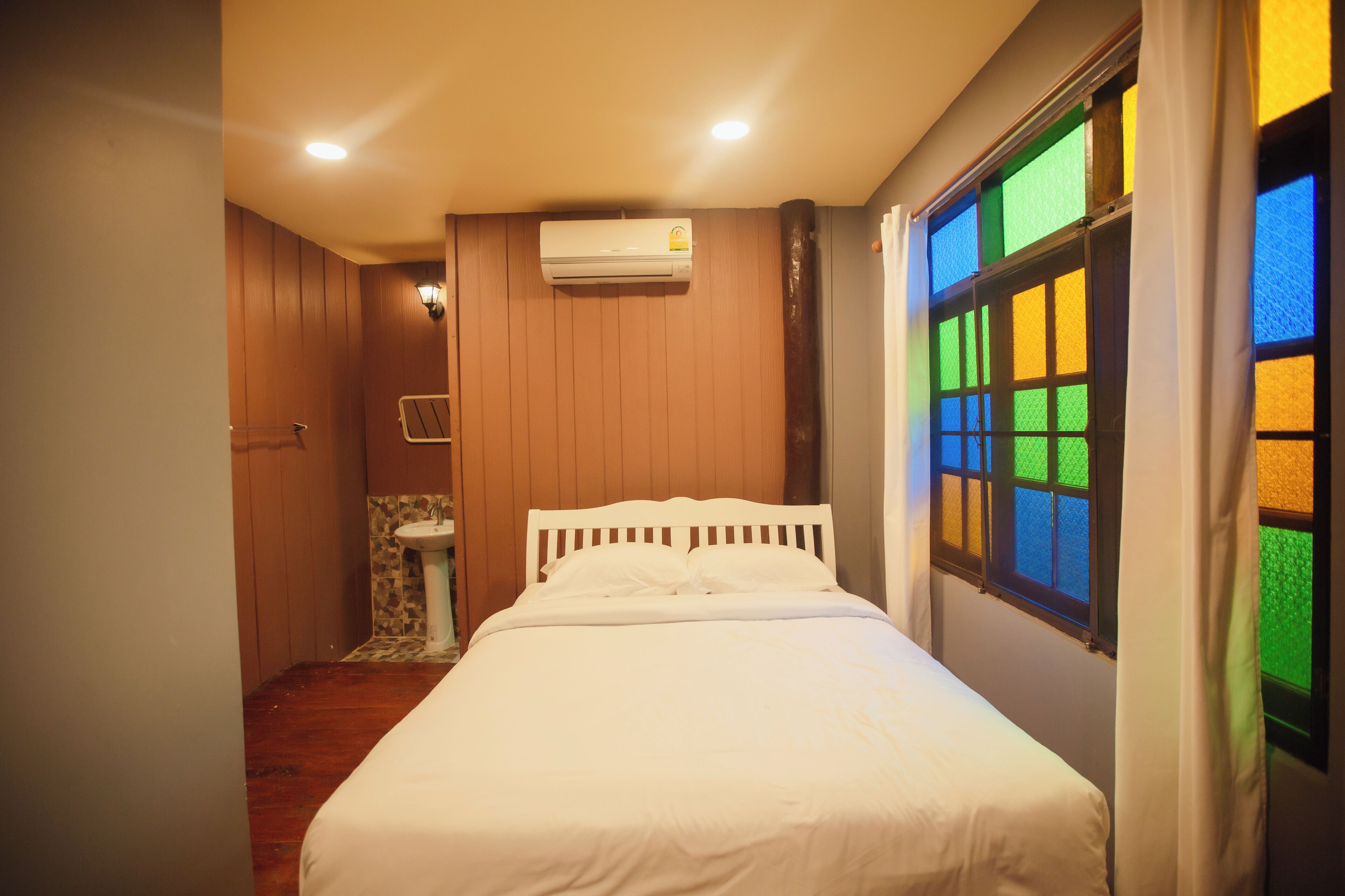 A bedroom with a bed, a window with colorful glass panes, a wall-mounted air conditioner, and a small bathroom with a sink and toilet.