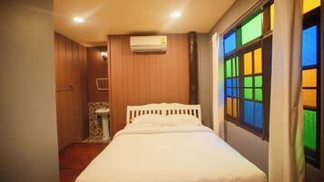 A bedroom with a bed, a window with colorful glass panes, a wall-mounted air conditioner, and a small bathroom with a sink and toilet.