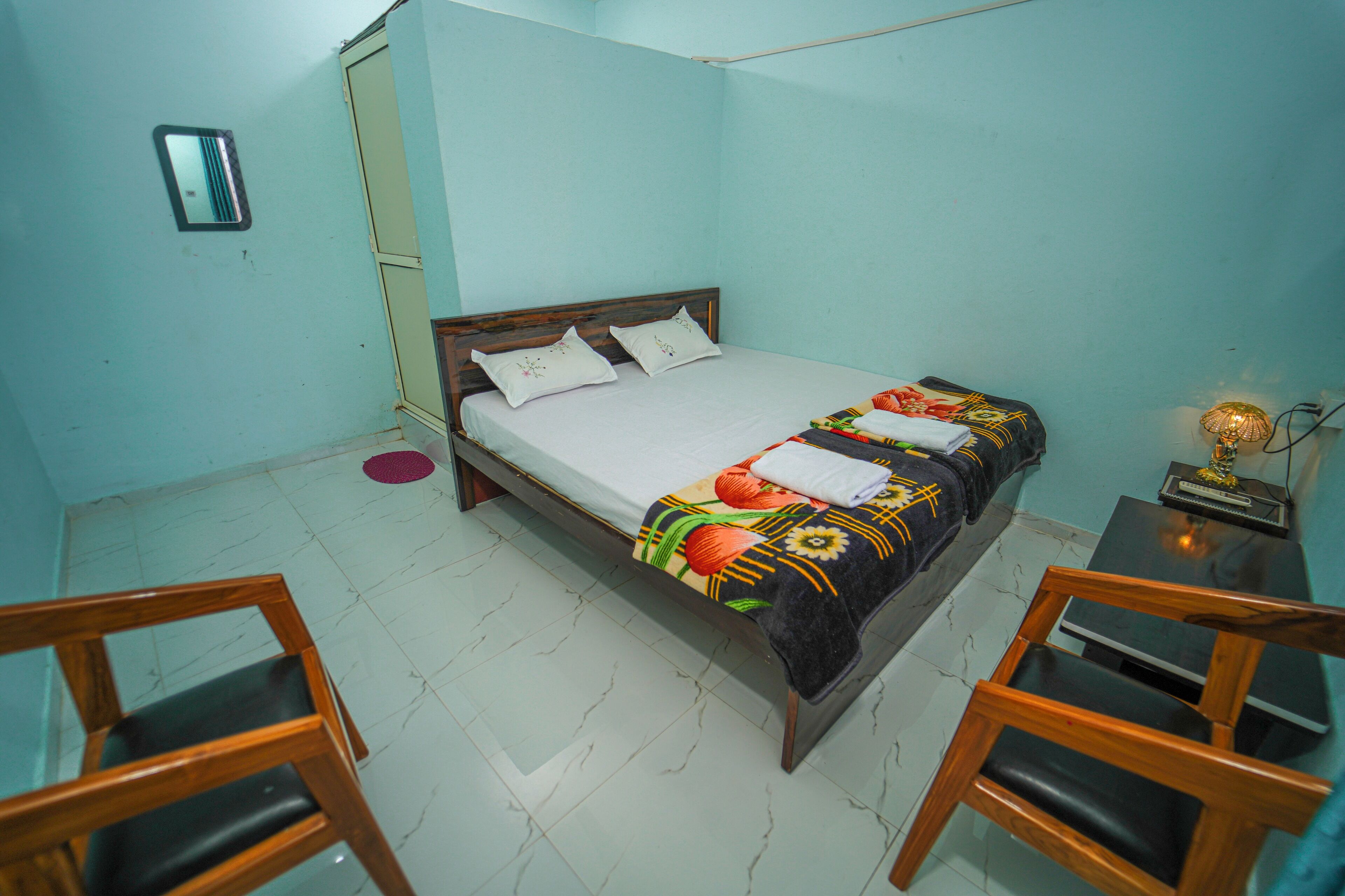 A small, clean room with a bed, two wooden chairs, a small table, and a wall-mounted mirror.