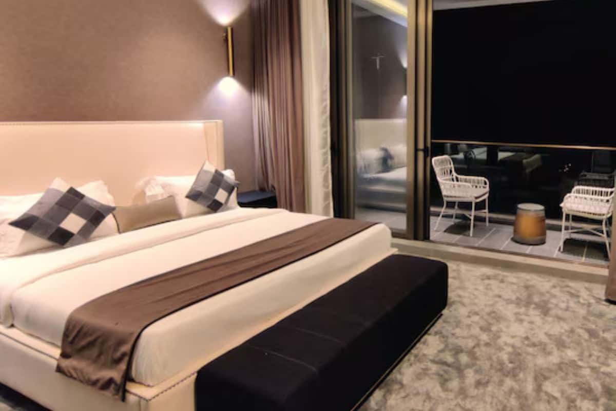 A modern hotel room with a large bed, a small seating area with chairs, and a balcony with a view.