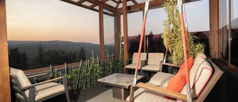 Zona de estar al aire libre iluminada por el sol con una silla colgante, una mesa y sillas, con vistas a un paisaje pintoresco.