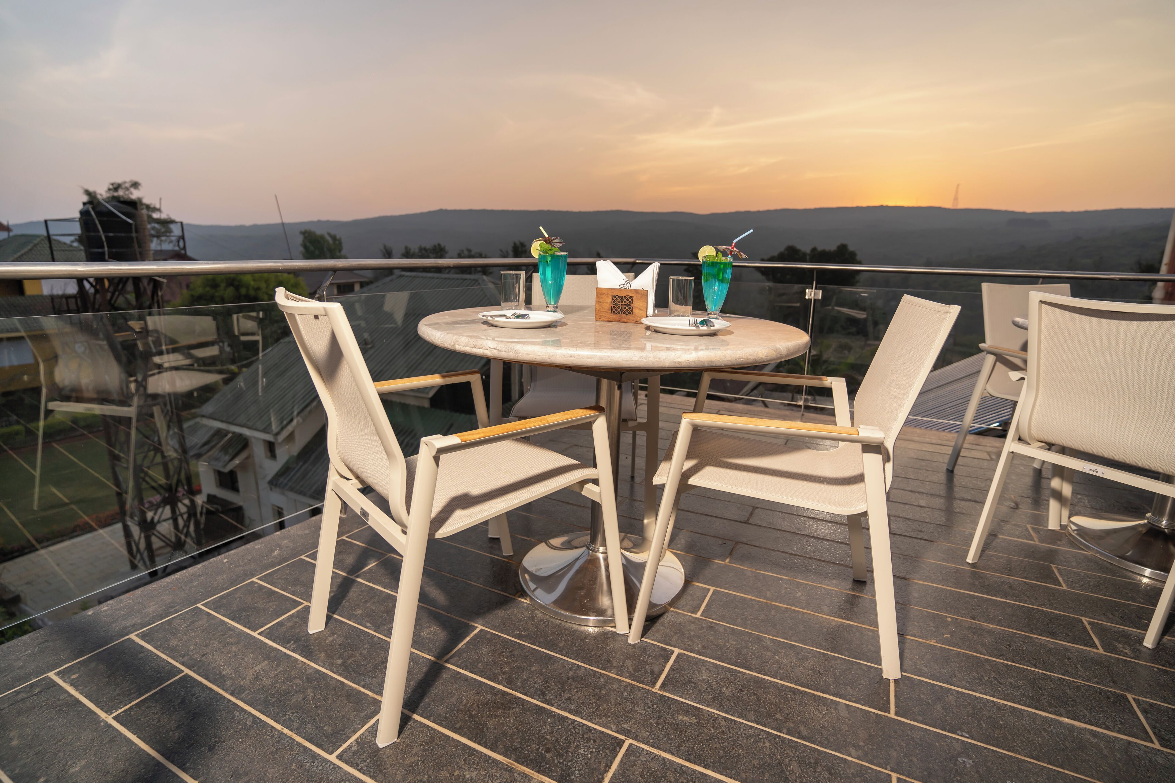 Una mesa redonda con cuatro sillas en un balcón con barandilla de vidrio, con vistas panorámicas a colinas y un atardecer.