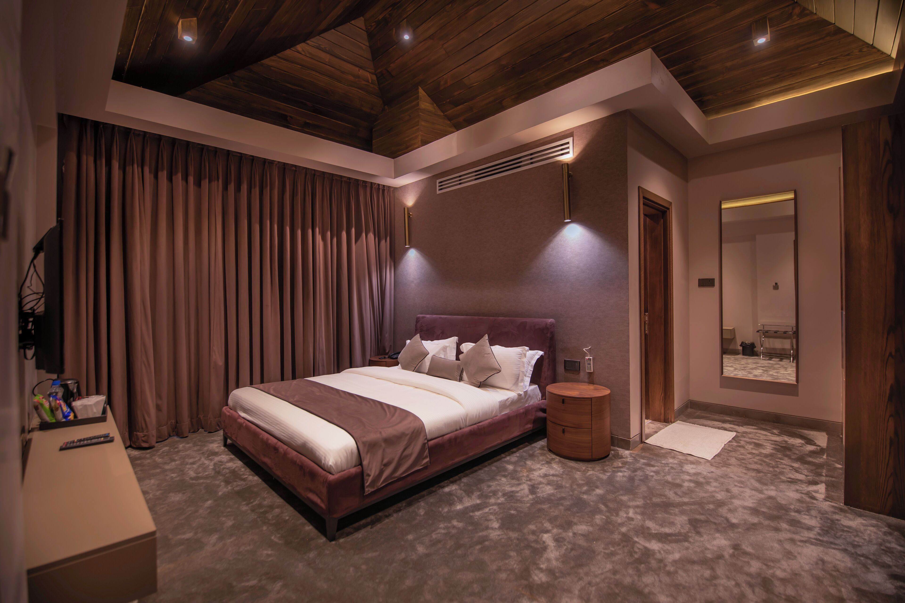 Un dormitorio con una cama grande, una mesita de noche, un tablero auxiliar, suelo de madera, techo con vigas de madera y un espejo instalado en la pared.