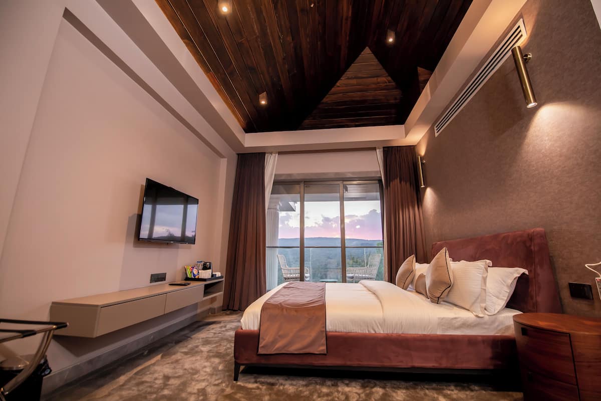 A modern bedroom with a large bed, a TV mounted on the wall, a desk with a chair, and a view of the outdoors through a large window.