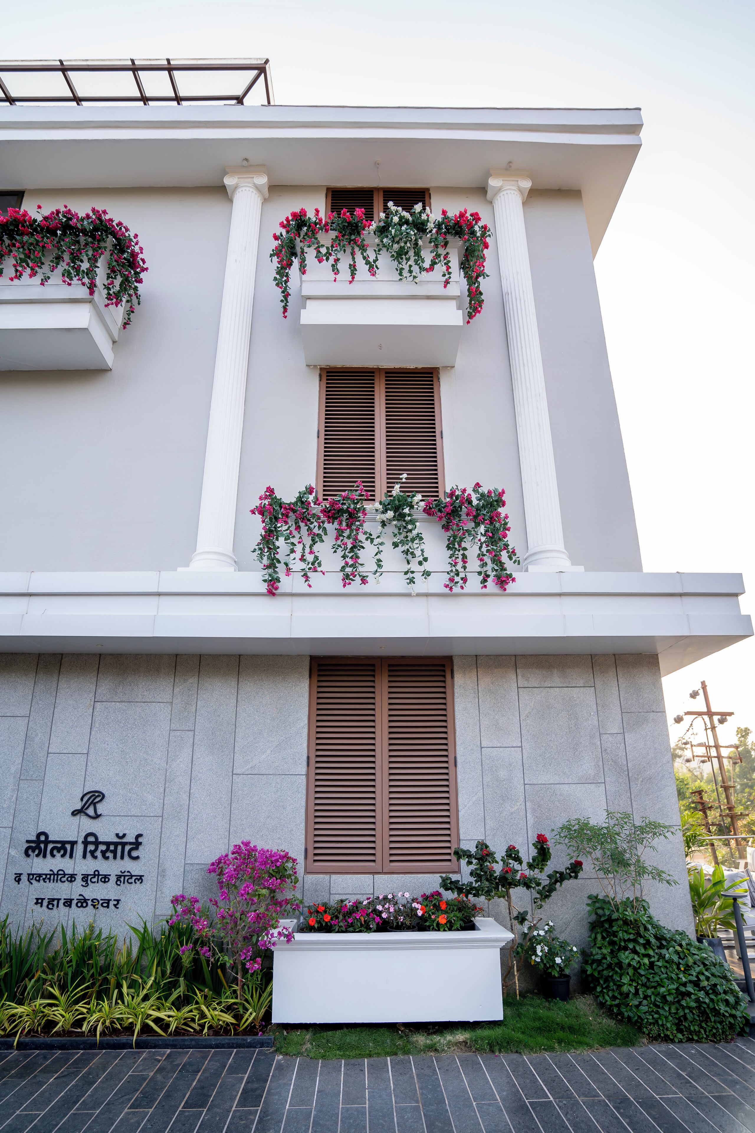 Um edifício com colunas brancas, uma varanda florida com flores cor-de-rosa e uma placa com texto.