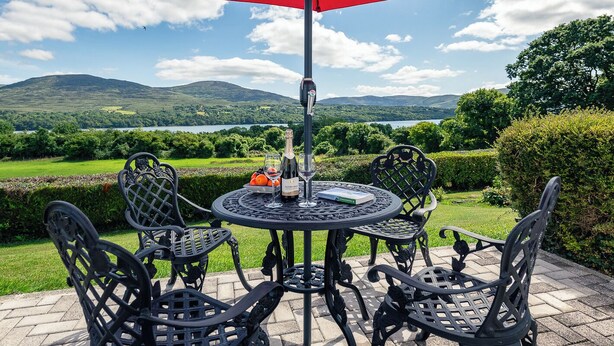 Een ronde tafel met vier stoelen en een rode parasol, op een terras met een prachtig uitzicht op heuvels en een meer.
