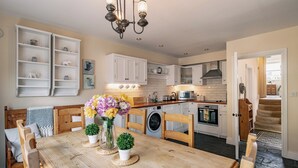 Eine moderne Küche mit weißen Schränken, einem Holztisch mit Stühlen, einer Kristallleuchter und Blick ins Wohnzimmer.