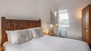 Un dormitorio con cabecera de madera, cama con ropa de cama blanca, mesita de noche con lámpara y ventana con vista a edificios.