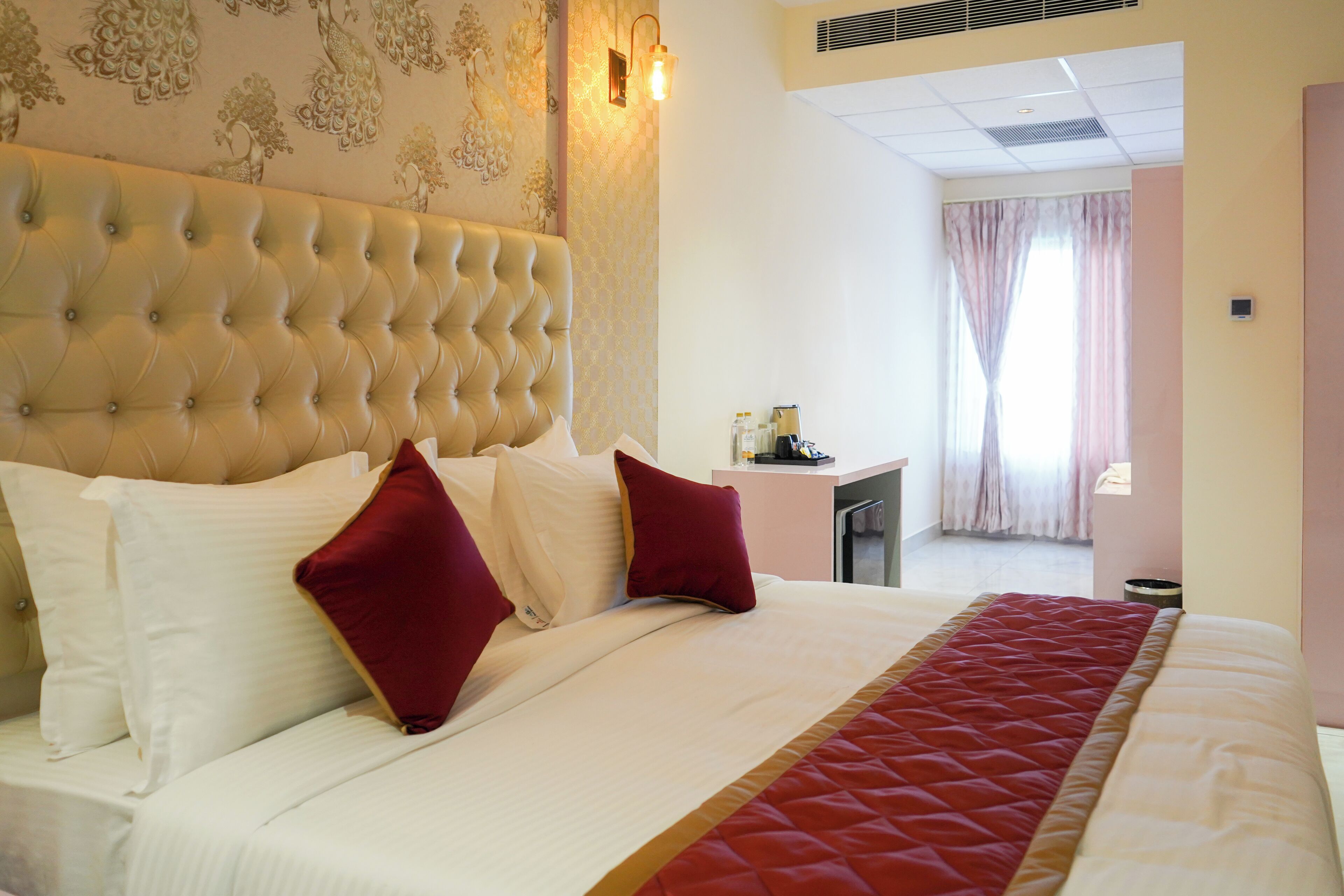 Quarto de hotel com cabeceira acolchoada, cama com roupa de cama branca e vermelha, uma pequena mesa de escritório com artigos de higiene pessoal e vista do quarto através de uma janela com cortinas.