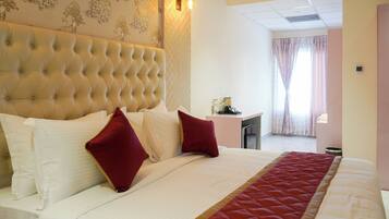 Quarto de hotel com cabeceira acolchoada, cama com roupa de cama branca e vermelha, uma pequena mesa de escritório com artigos de higiene pessoal e vista do quarto através de uma janela com cortinas.