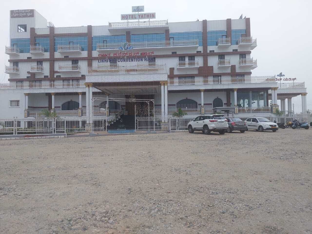 A multi-story hotel with a central entrance, balconies, and a parking area with several cars.
