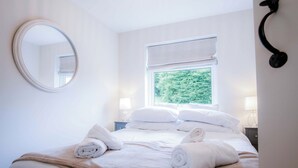 A neatly made bed with white linens and rolled towels, a round mirror on the wall, and a window with a view of greenery. - Bayview 3 - 2 Bedroom - Bayview Oxwich (Swansea)