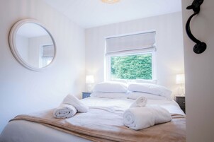 Una cama bien hecha con ropa de cama blanca y toallas enrolladas, un espejo redondo en la pared y una ventana con vistas a zonas verdes.