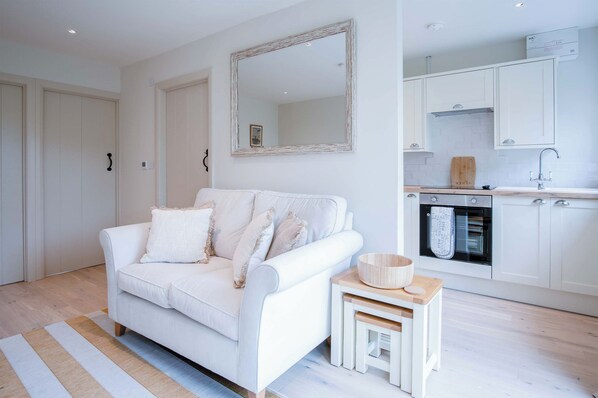A white living room with a sofa, a small wooden side table, and a kitchen area with white cabinets and a built-in oven.