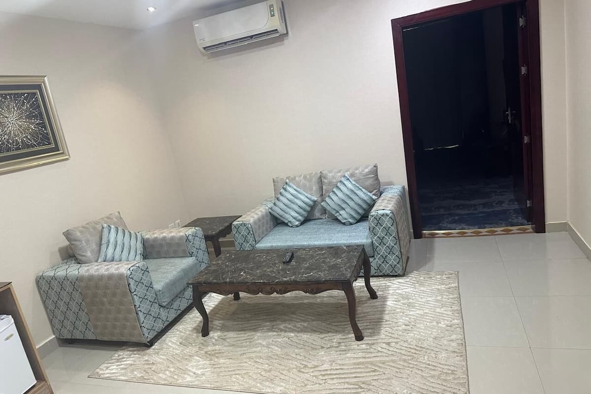 A living room with a sofa, armchair, and coffee table. There is an air conditioning unit on the wall and a door leading to another room.