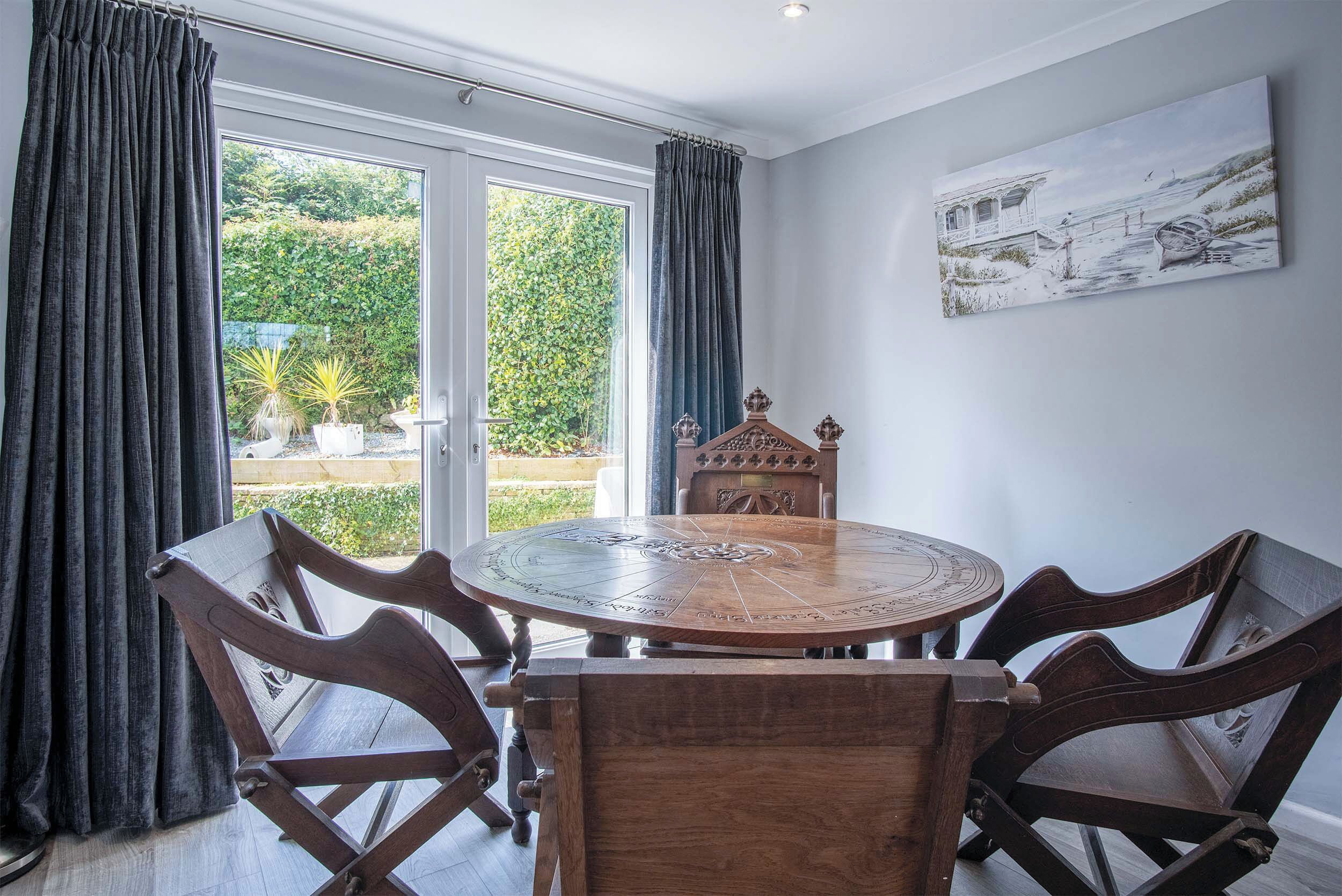 A dining area with a round wooden table and chairs, a painting on the wall, and a view through the window.