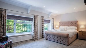 A bedroom with a large window, a tufted headboard, a bedside table with a lamp, and a chair with a patterned cushion. - Bryn View – 5 Bedroom Holiday Home – Oxwich (Swansea)