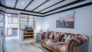 A modern kitchen with wooden cabinets and a built-in oven, a cozy living area with a large, tufted sofa, and a view of the sea through a large window. - Bryn View – 5 Bedroom Holiday Home – Oxwich (Swansea)
