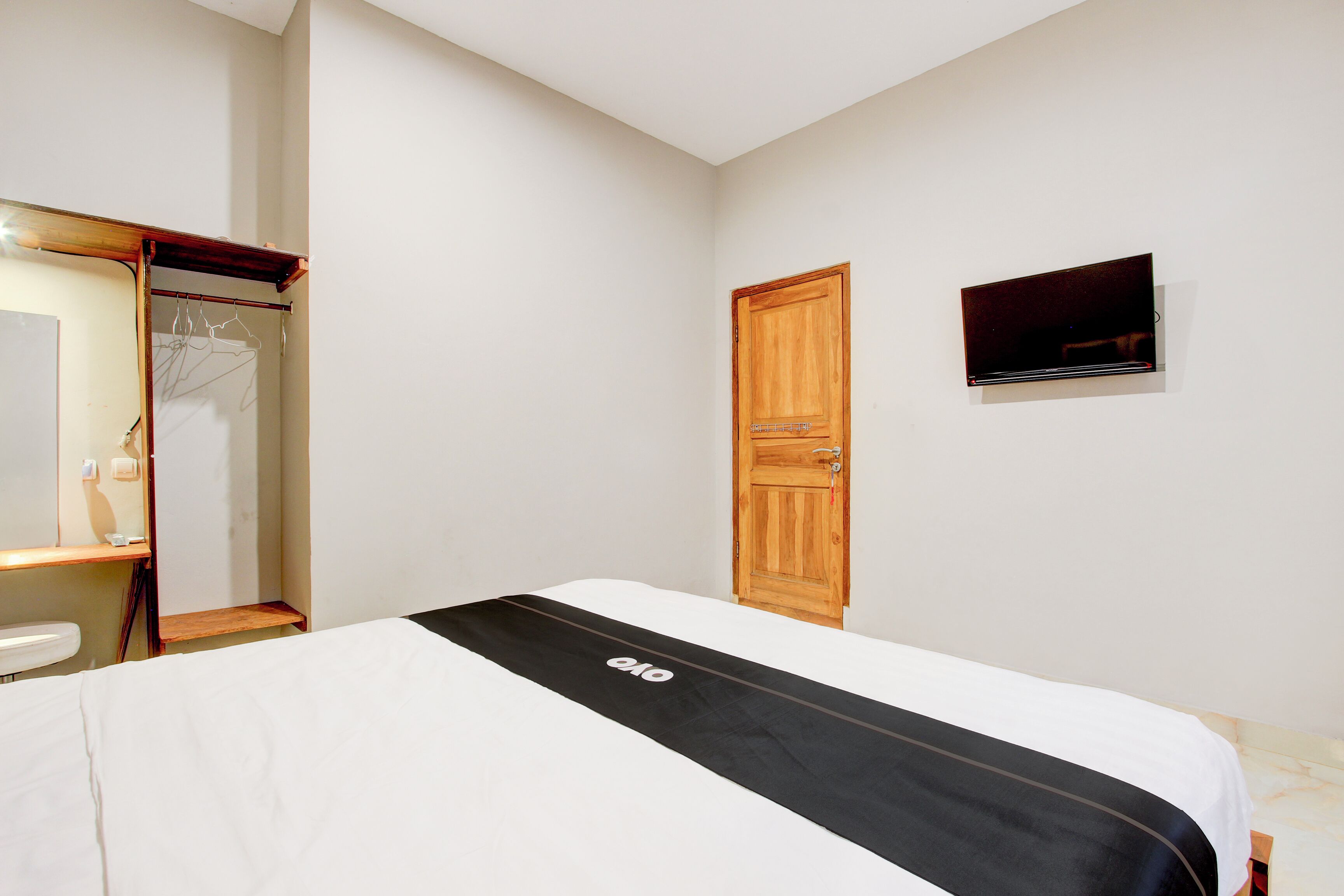 A bedroom with a bed, a wooden wardrobe, a mirror, and a television mounted on the wall.