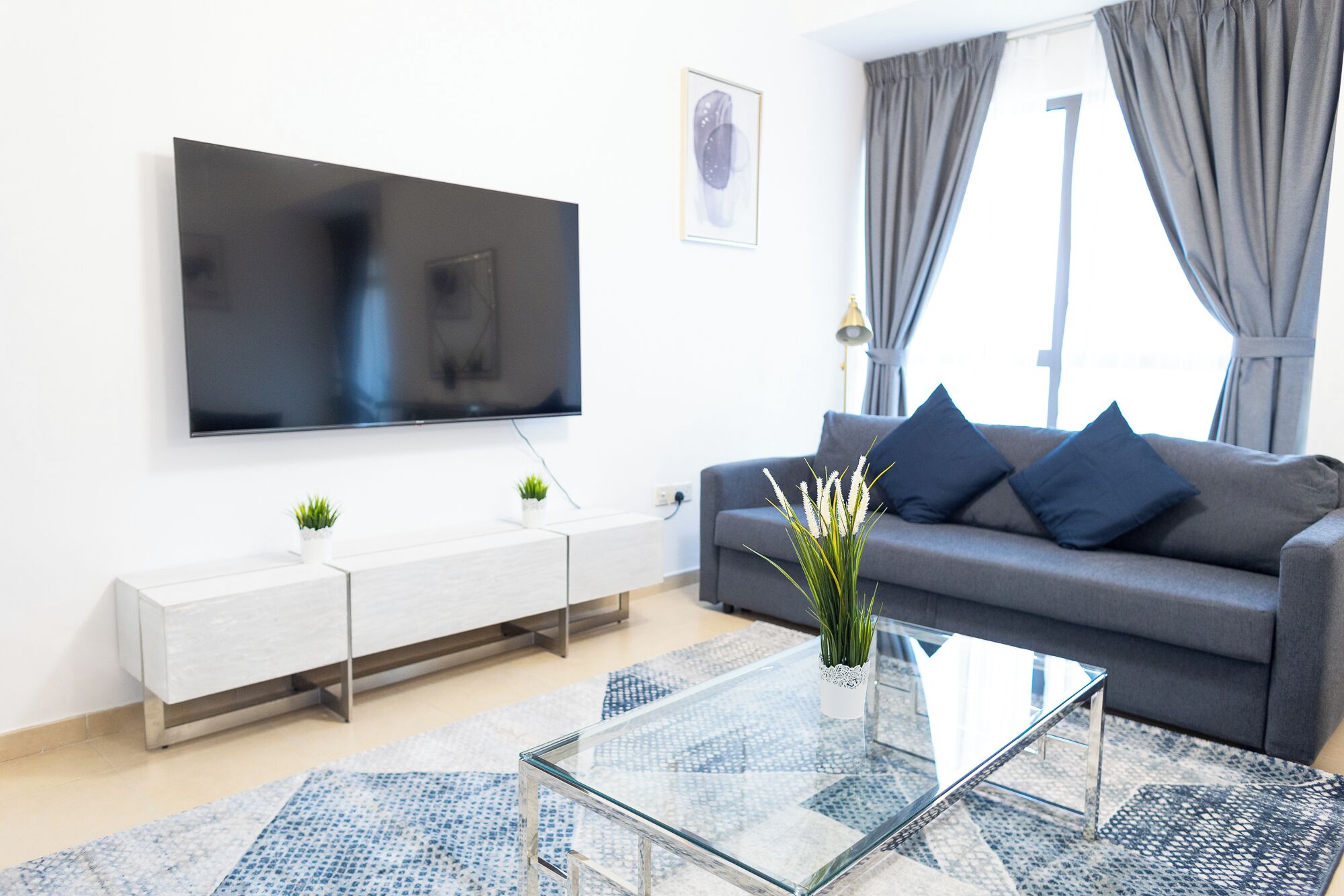 Egy modern nappali, melyben a falra van szerelve egy síkképernyős televízió, egy szürke kanapé kék párnákkal, egy üveg kávézóasztal és egy mintás szőnyeg található.