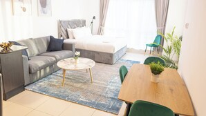 Et moderne hotelværelse med en grå sofa, en seng med gråt sengegavl, et rundt sofabord, et træspisebord med stole og et stort vindue med gardiner.