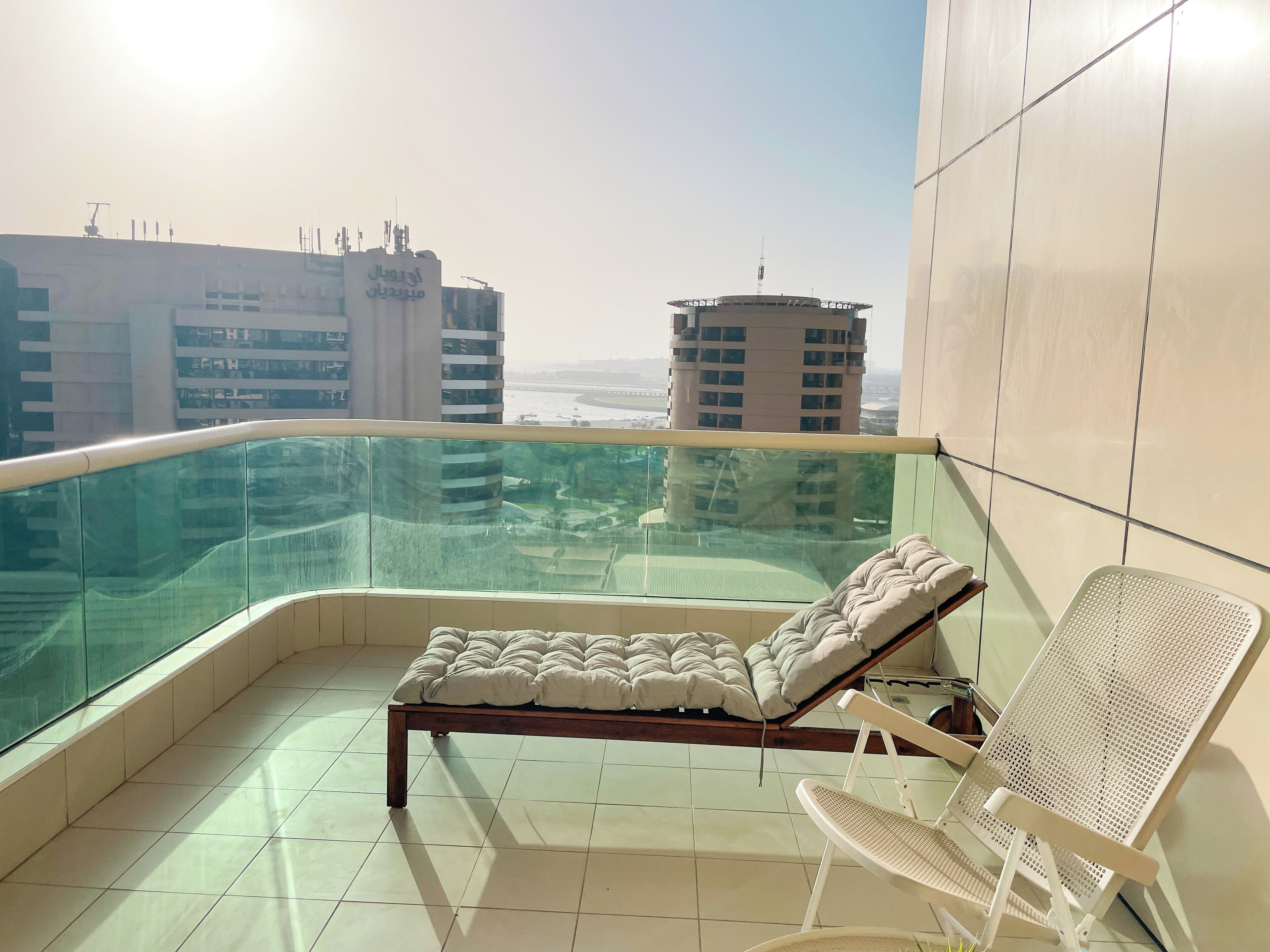 A balcony with a bed and a chair, overlooking a cityscape with tall buildings.