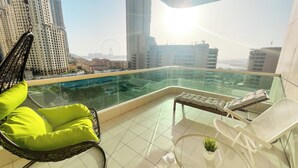 En balkon med gelænder af glas, en fletstol med grøn pude, et lille bord med en plante og udsigt over en by med høje bygninger og et pariserhjul.