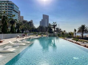 A swimming pool with lounge chairs, palm trees, and modern buildings in the background. - HAPPY SEASON I La Vie apartment  (Dubai)