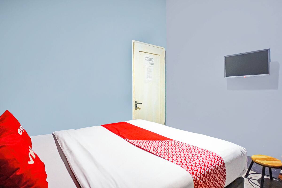 A room with a bed, a door, a small stool, and a flat-screen TV mounted on the wall.