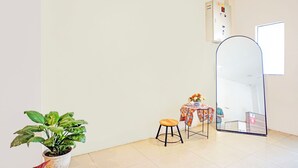 Een kamer met een potplant, een klein krukje, een tafel met een kleurrijk tafelkleed en een grote spiegel.