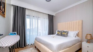 Une chambre d’hôtel avec un grand lit, un fer à repasser et une planche à repasser, une table de chevet et une vue sur la ville par la fenêtre.