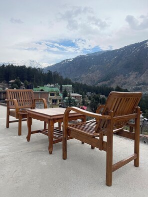 A wooden table and chairs set on a concrete surface with a mountain view in the background. - Shubham Cottage (Manali)