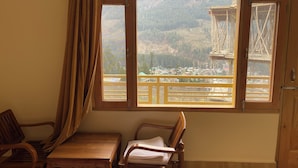 A room with a large window overlooking a mountainous landscape and a construction site. There are two wooden chairs and a small table. - Shubham Cottage (Manali)