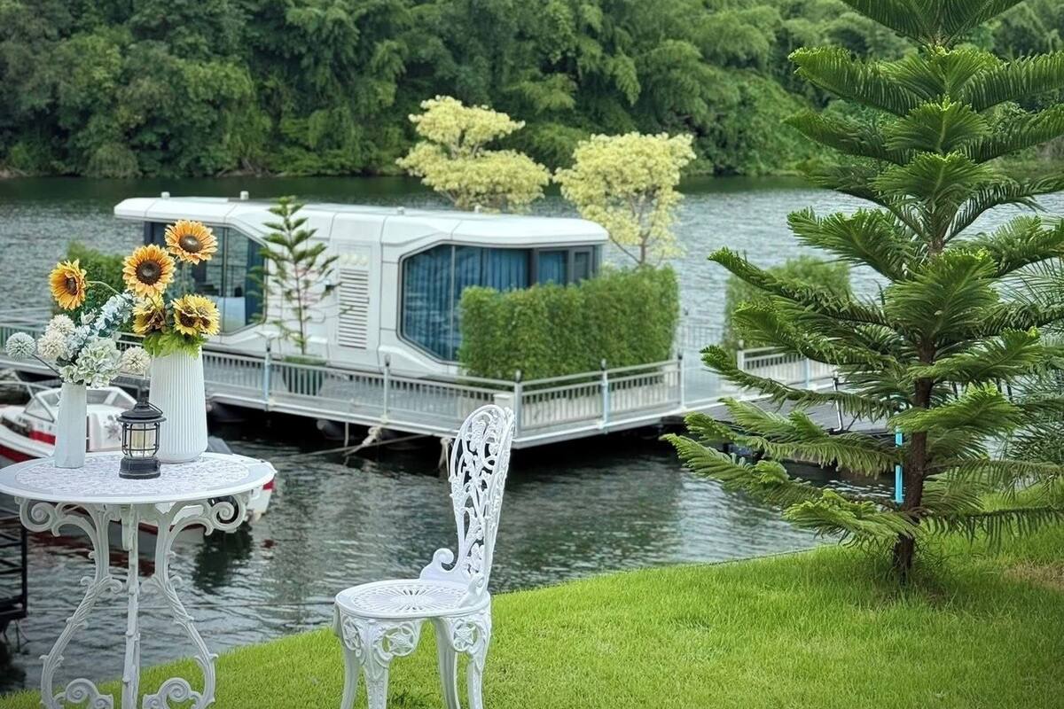 A lakeside setting with a white boat docked near a grassy area, a white table with a vase of sunflowers, and a white chair.