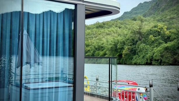 Una cabaña moderna con un muro de cristal, un área de terraza con sombrillas de colores y vistas a un lago y montañas boscosas.