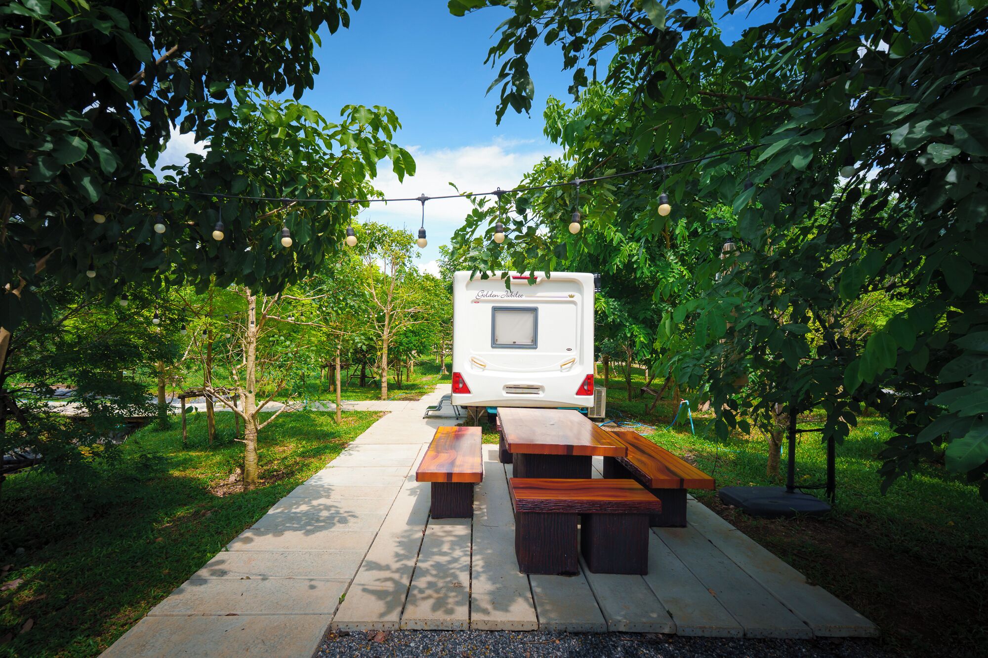 Una caravana blanca estacionada en un jardín exuberante, con mesas de picnic y luces colgantes.