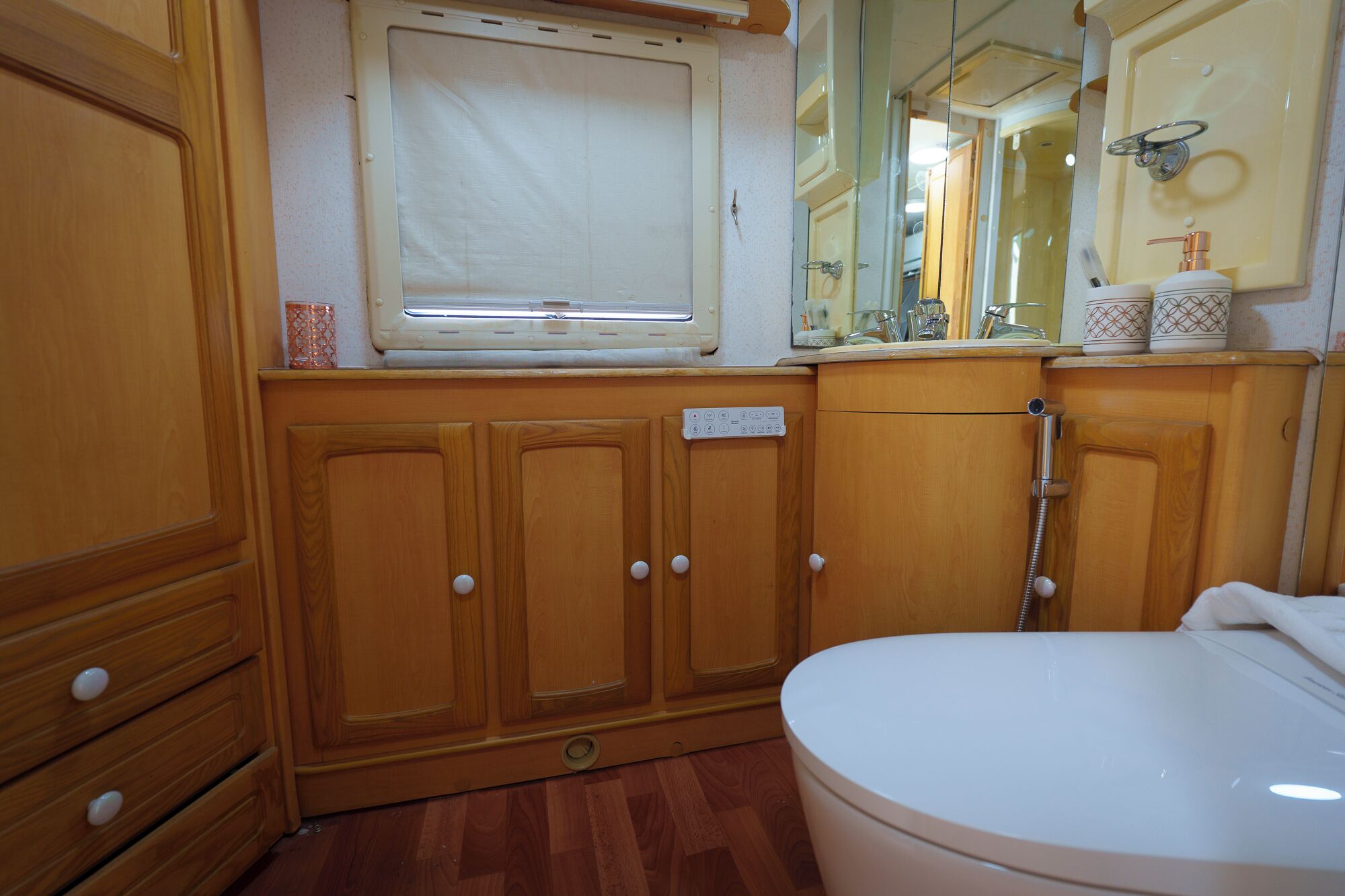 A compact bathroom with wooden cabinetry, a toilet, a window, and a mirror.