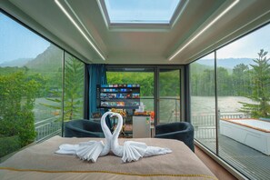 A modern room with a large window overlooking a river and greenery, a bed with a swan towel arrangement, and a flat-screen TV on a stand.
