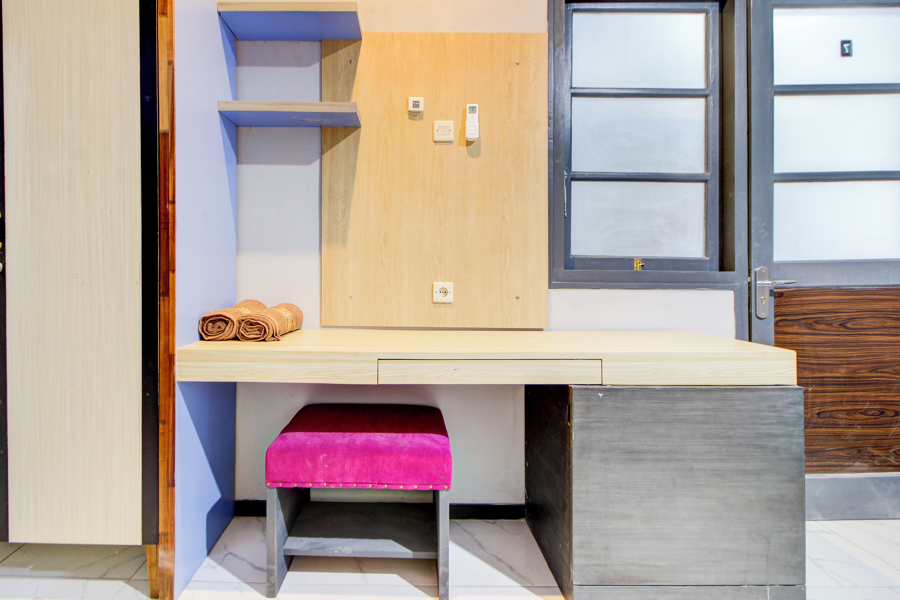 Una habitación compacta con un escritorio de madera, un banco rosa y un ventanal con paneles de vidrio esmerilado.