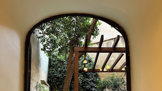 A view through an arched window of a swimming pool area with a wooden pergola, potted plants, and a small deck.